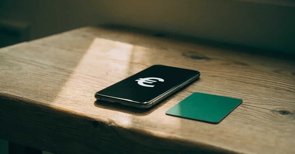 Ein Smartphone liegt mit dem Display nach unten auf einem Holztisch, daneben ein paar Euro-Münzen und eine unbeschriftete Plastikkarte im Morgenlicht.